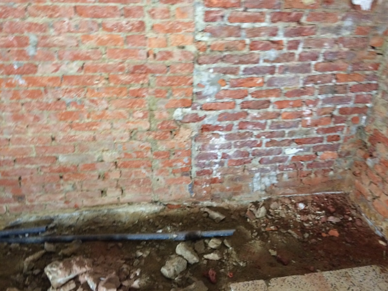 Blootgestelde rode bakstenen muur met wat puin en vuil op de vloer bij de hoek: ideaal voor een vochtbestrijdingsexpert of een specialist in waterdichting en betoninjectie om vochtproblemen te beoordelen en op te lossen.